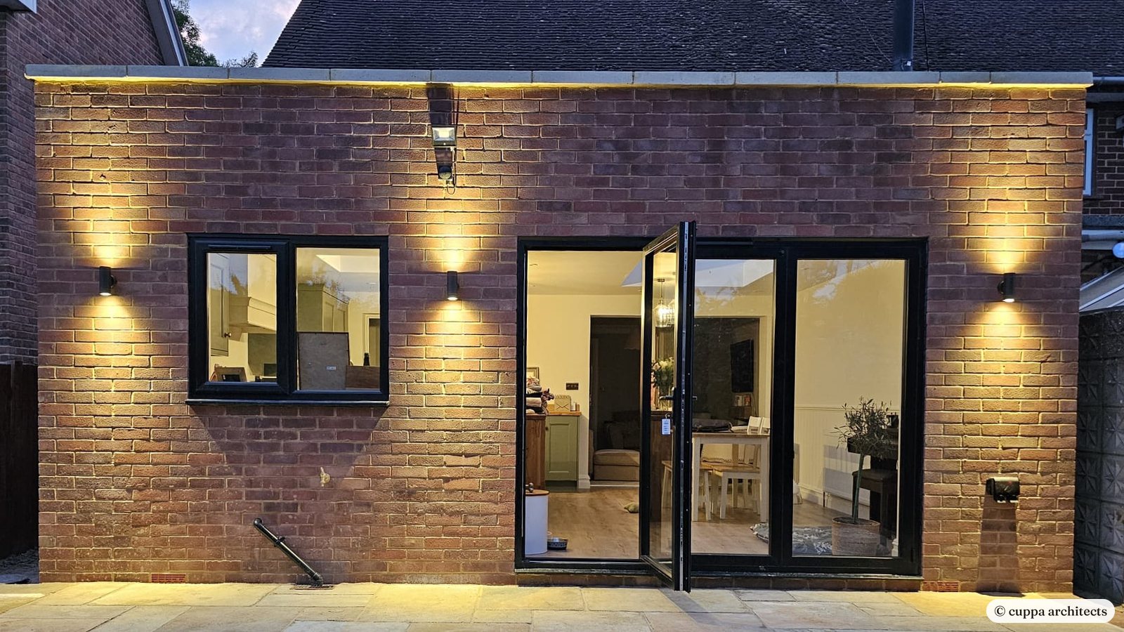 A brick rear extension lit up in the evening, with glazed doors to a stone terrace