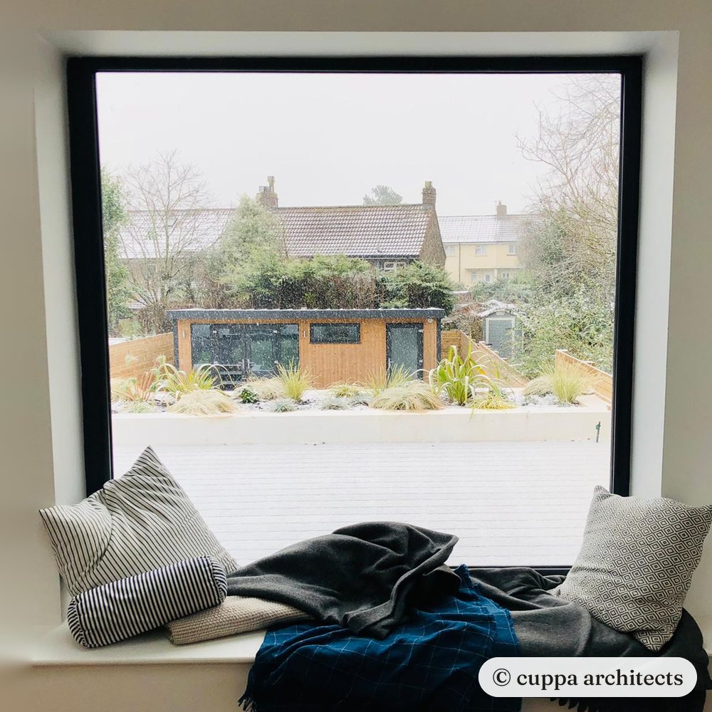 A window seat with cushions and throws, looking out over a snowy garden to a timber garden studio